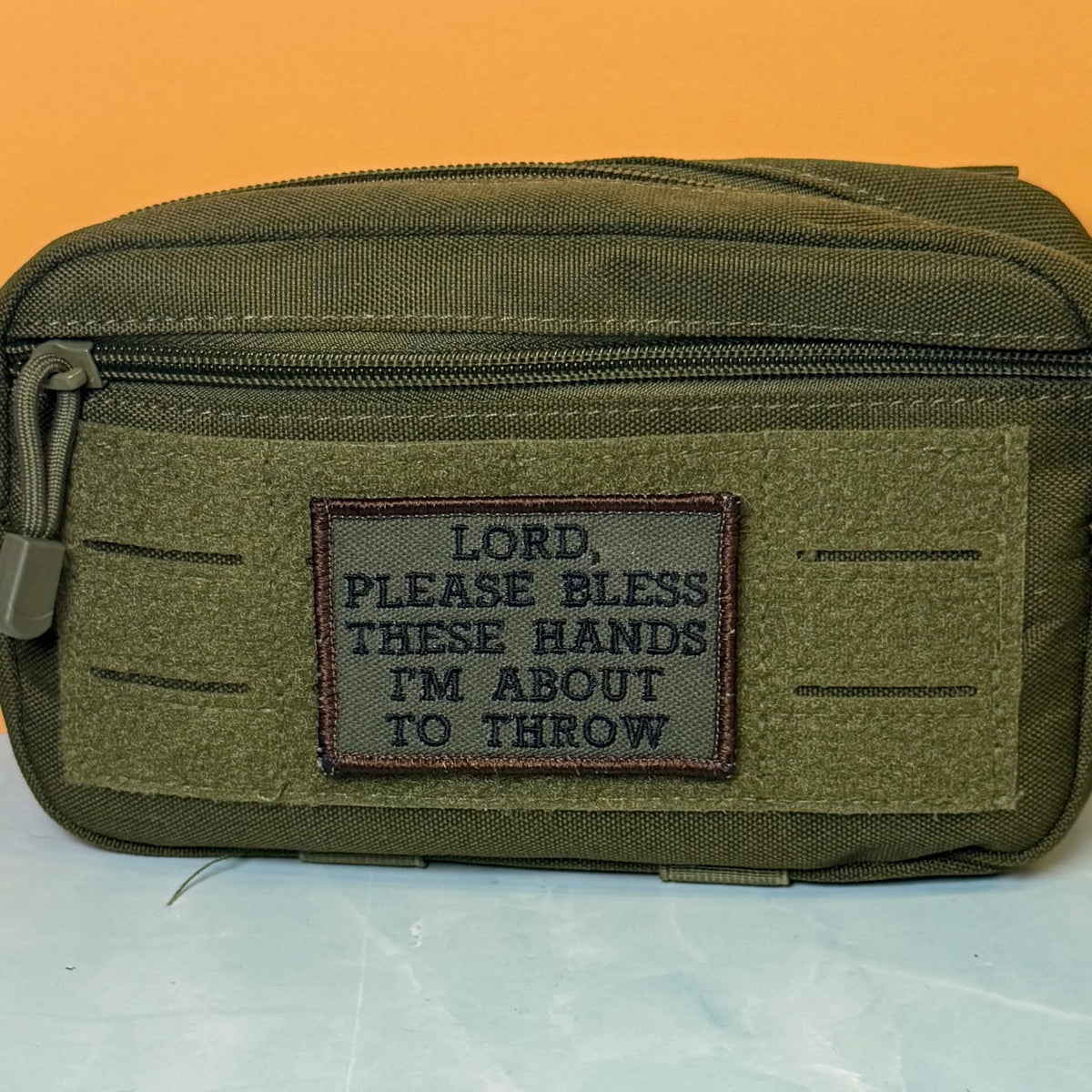 Lord, Bless These Hands I'm About To Throw embroidered morale patch: olive green tactical pouch with a front hook-and-loop patch that reads, 'LORD, PLEASE BLESS THESE HANDS I'M ABOUT TO THROW,' shown against an orange.