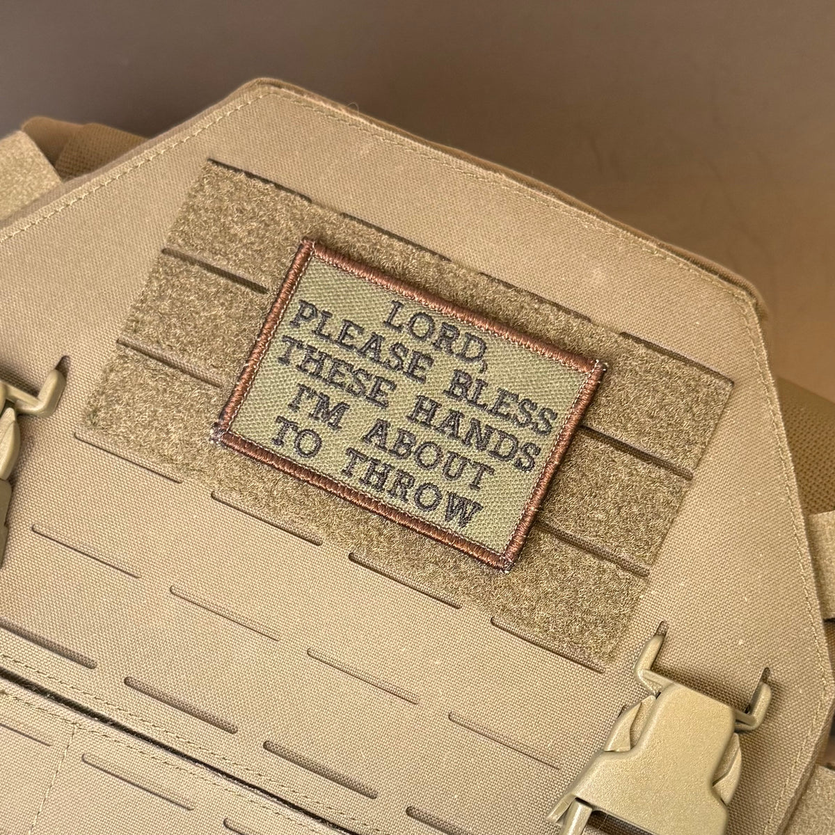 Lord, Bless These Hands I'm About To Throw embroidered morale patch: beige tactical bag with a rectangular embroidered patch reading 'LORD, PLEASE BLESS THESE HANDS I'M ABOUT TO THROW' on a tan webbing panel.