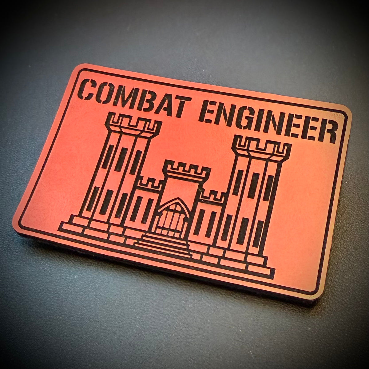 Combat Engineer Army Veteran laser-engraved synthetic patch: a rectangular orange-red patch or patch with black border, black text reading 'COMBAT ENGINEER,' and a black line drawing of a fort or castle, photographed on a.
