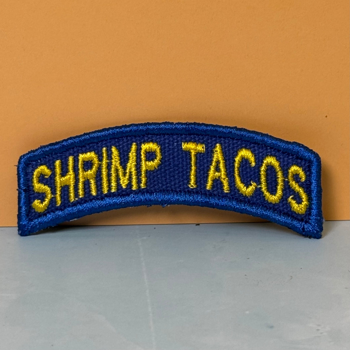 Army Regular Forces Funny Morale Patch Tab embroidered morale patch: curved patch with the words 'SHRIMP TACOS' in yellow lettering on a blue background, placed against a solid orange wall and gray surface.