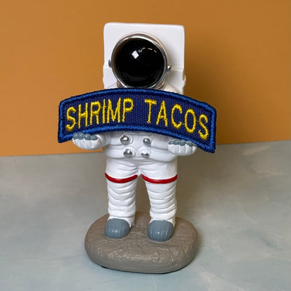 Army Regular Forces Funny Morale Patch Tab embroidered morale patch: astronaut figurine holding a blue embroidered patch that reads 'SHRIMP TACOS' in yellow lettering, standing on a gray base against an orange and gray background.