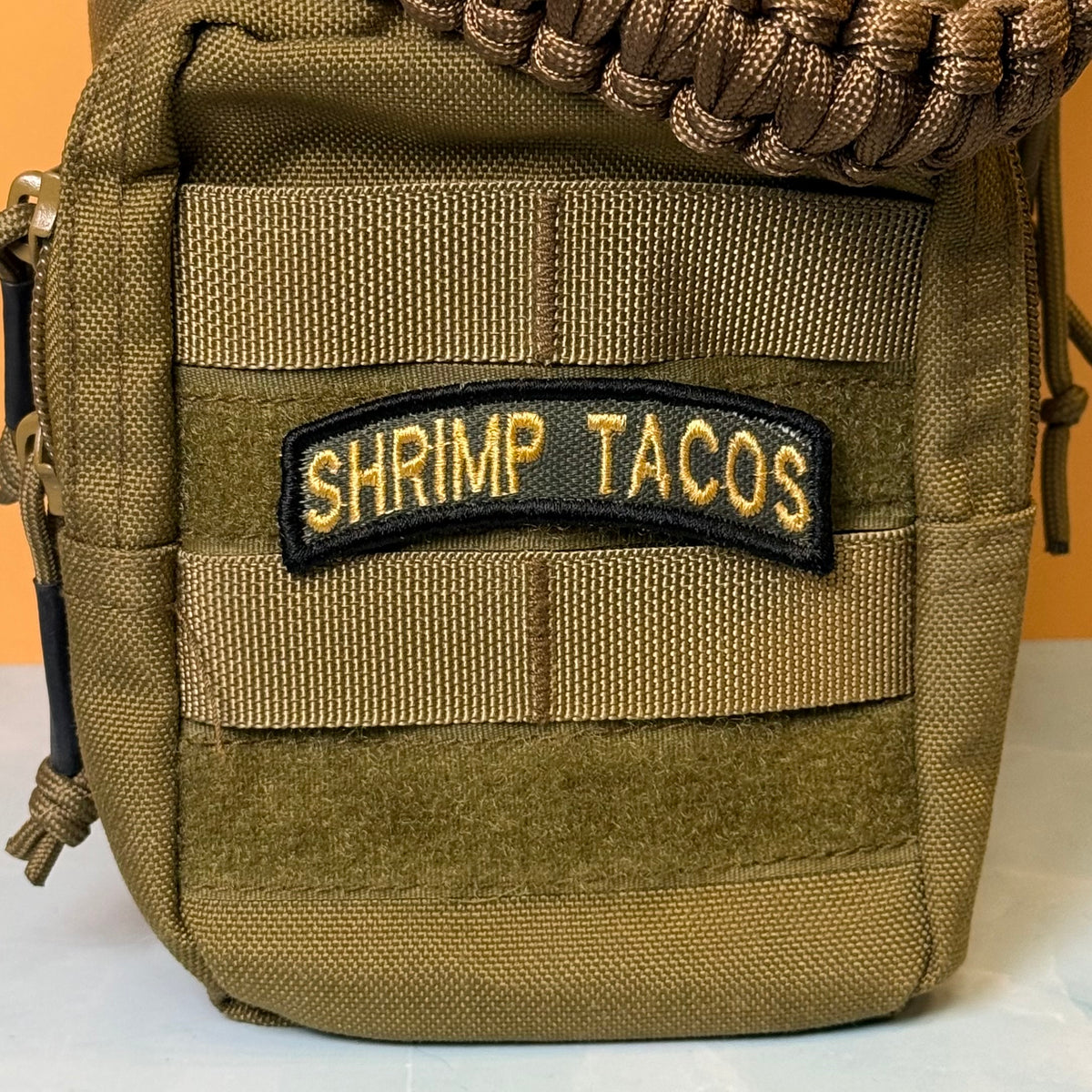 Army Regular Forces Funny Morale Patch Tab embroidered morale patch: olive drab tactical pouch with MOLLE webbing and a black embroidered patch reading 'SHRIMP TACOS' in gold lettering, shown against a solid orange background.