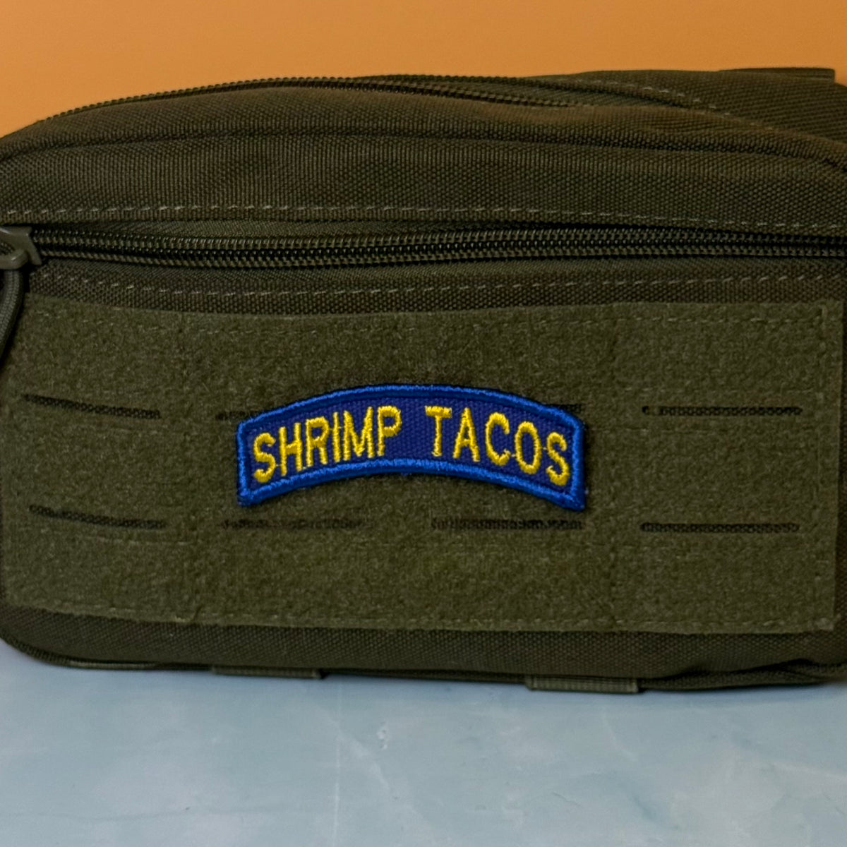 Army Regular Forces Funny Morale Patch Tab embroidered morale patch: olive drab green tactical pouch with a front zipper and a blue and yellow embroidered patch reading 'SHRIMP TACOS' on a light blue surface against an orange background.