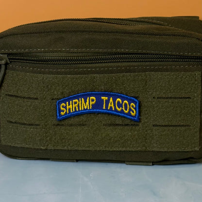 Army Regular Forces Funny Morale Patch Tab embroidered morale patch: olive drab green tactical pouch with a front zipper and a blue and yellow embroidered patch reading 'SHRIMP TACOS' on a light blue surface against an orange background.