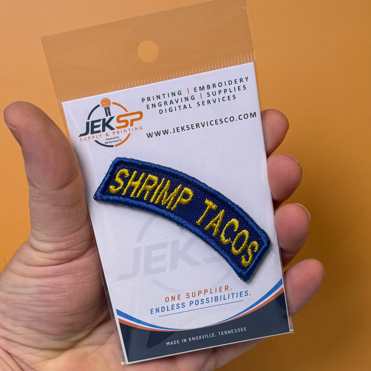 Army Regular Forces Funny Morale Patch Tab embroidered morale patch: hand holds a packaged embroidered patch that reads “SHRIMP TACOS” in yellow letters on a dark blue curved patch, mounted on a white backing card with the JEK SP logo.