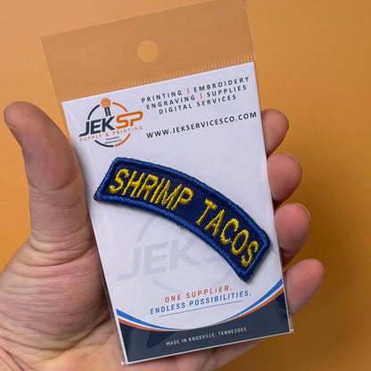 Army Regular Forces Funny Morale Patch Tab embroidered morale patch: hand holds a packaged embroidered patch that reads “SHRIMP TACOS” in yellow letters on a dark blue curved patch, mounted on a white backing card with the JEK SP logo.