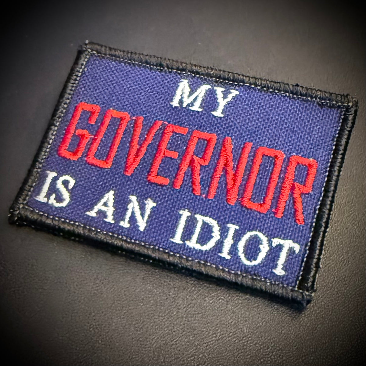 An embroidered rectangular patch with a dark blue background and black border, featuring the text 'MY GOVERNOR IS AN IDIOT' in red and white lettering, shown on a dark textured surface.