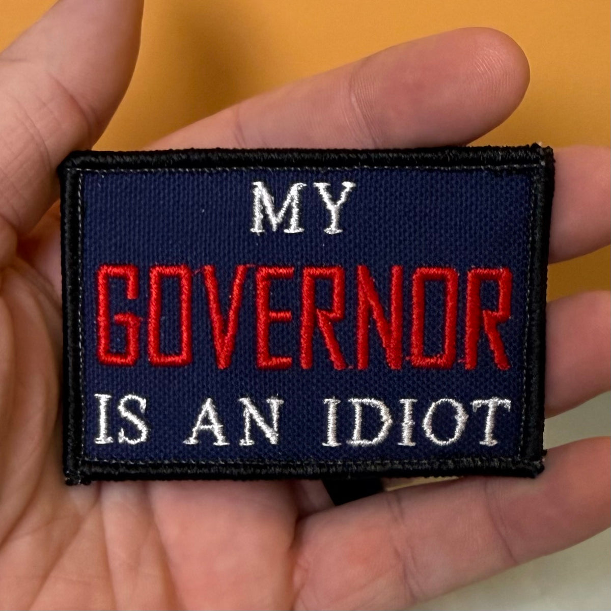 My Governor Is An Idiot embroidered morale patch: held in a hand, featuring the text 'MY GOVERNOR IS AN IDIOT' in red and white lettering on a dark navy blue background with a black stitched border.