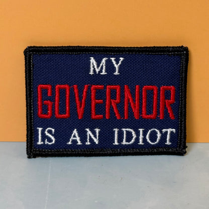 My Governor Is An Idiot embroidered morale patch: rectangular patch with the text 'MY GOVERNOR IS AN IDIOT' in white and red on a dark navy background, bordered in black, shown against an orange and gray background.