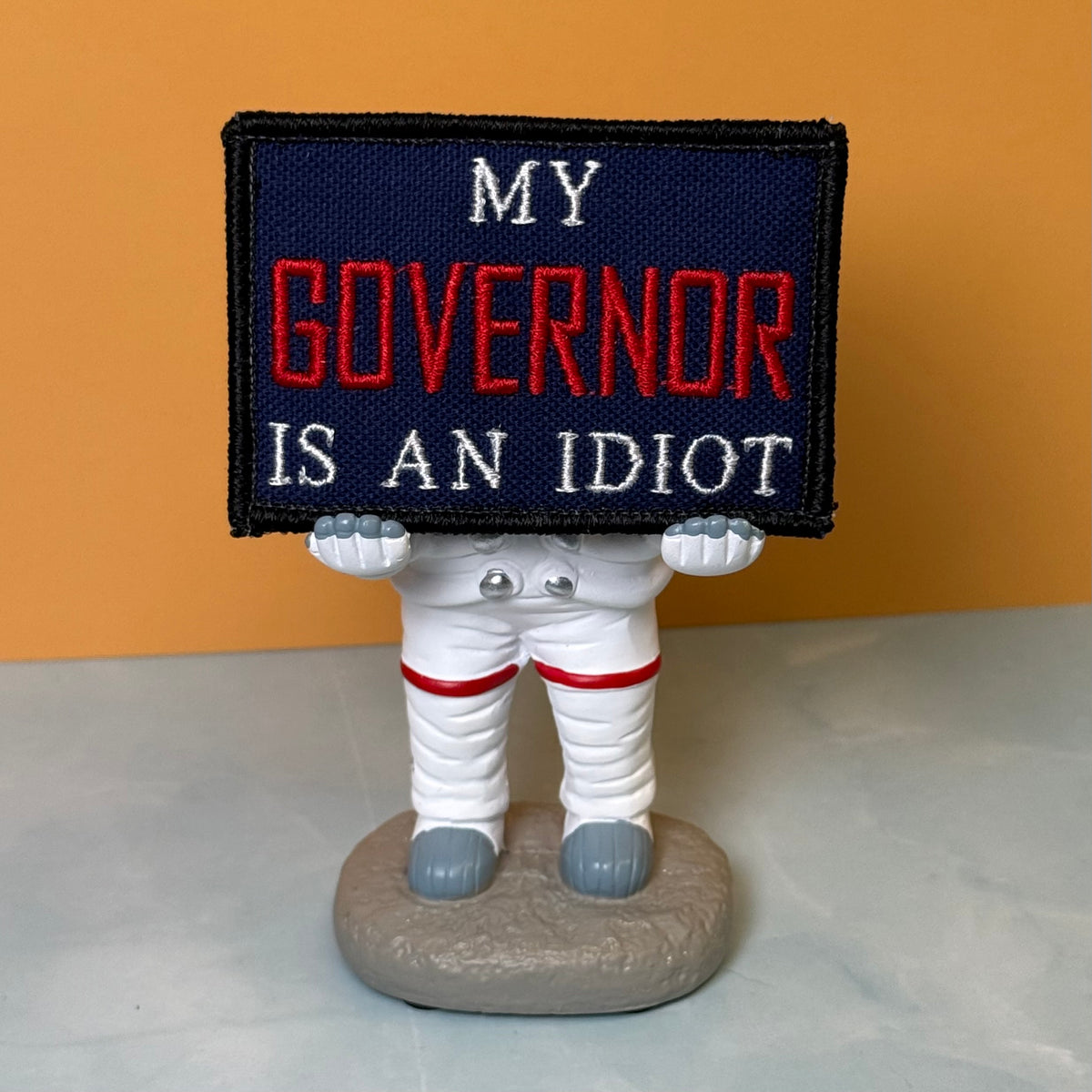 My Governor Is An Idiot embroidered morale patch: astronaut figurine holding a rectangular embroidered patch that reads 'MY GOVERNOR IS AN IDIOT' in white and red text on a dark navy background. The figurine is standing on a.
