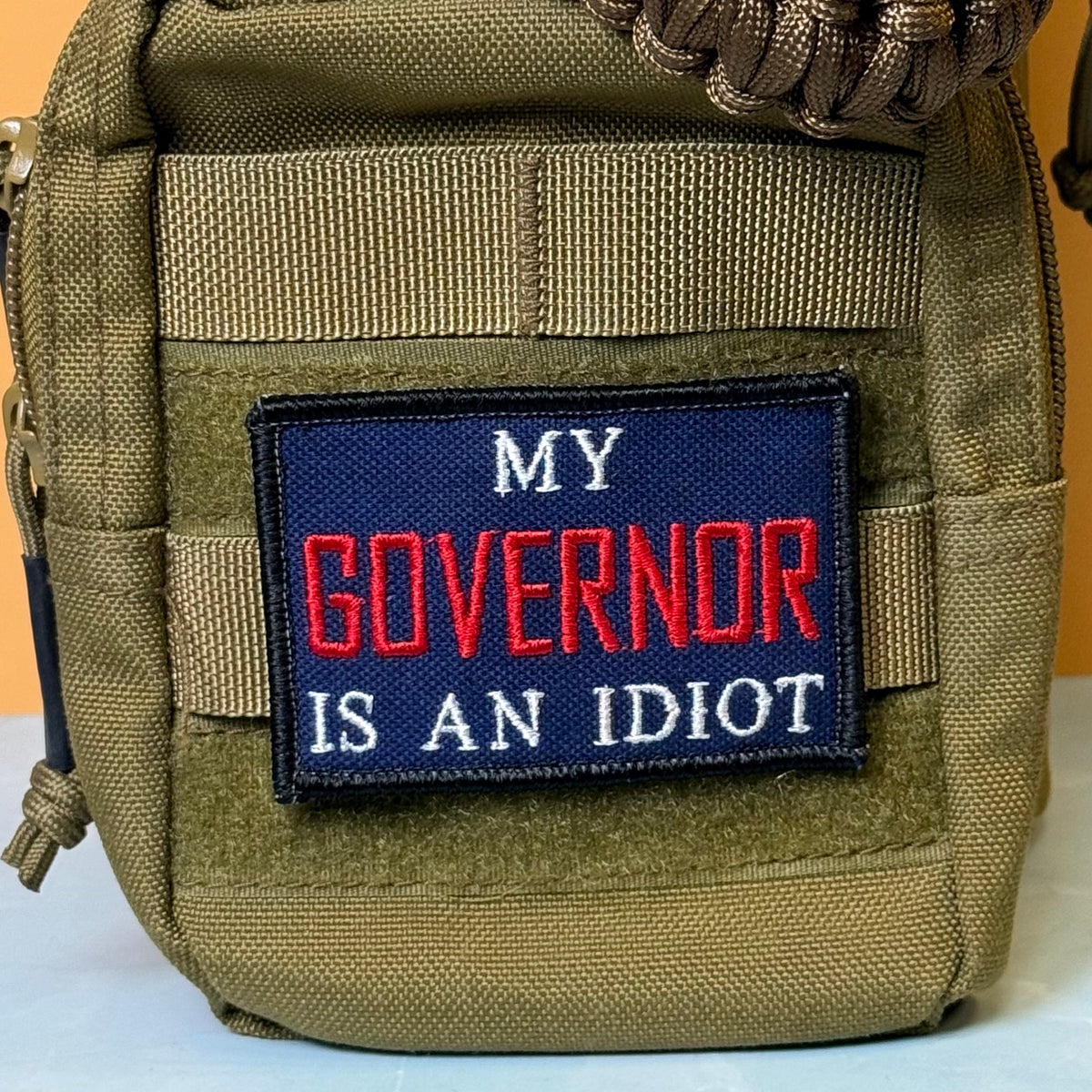 My Governor Is An Idiot embroidered morale patch: olive green tactical pouch featuring a rectangular embroidered patch with the text 'MY GOVERNOR IS AN IDIOT' in red and white on a dark blue background.