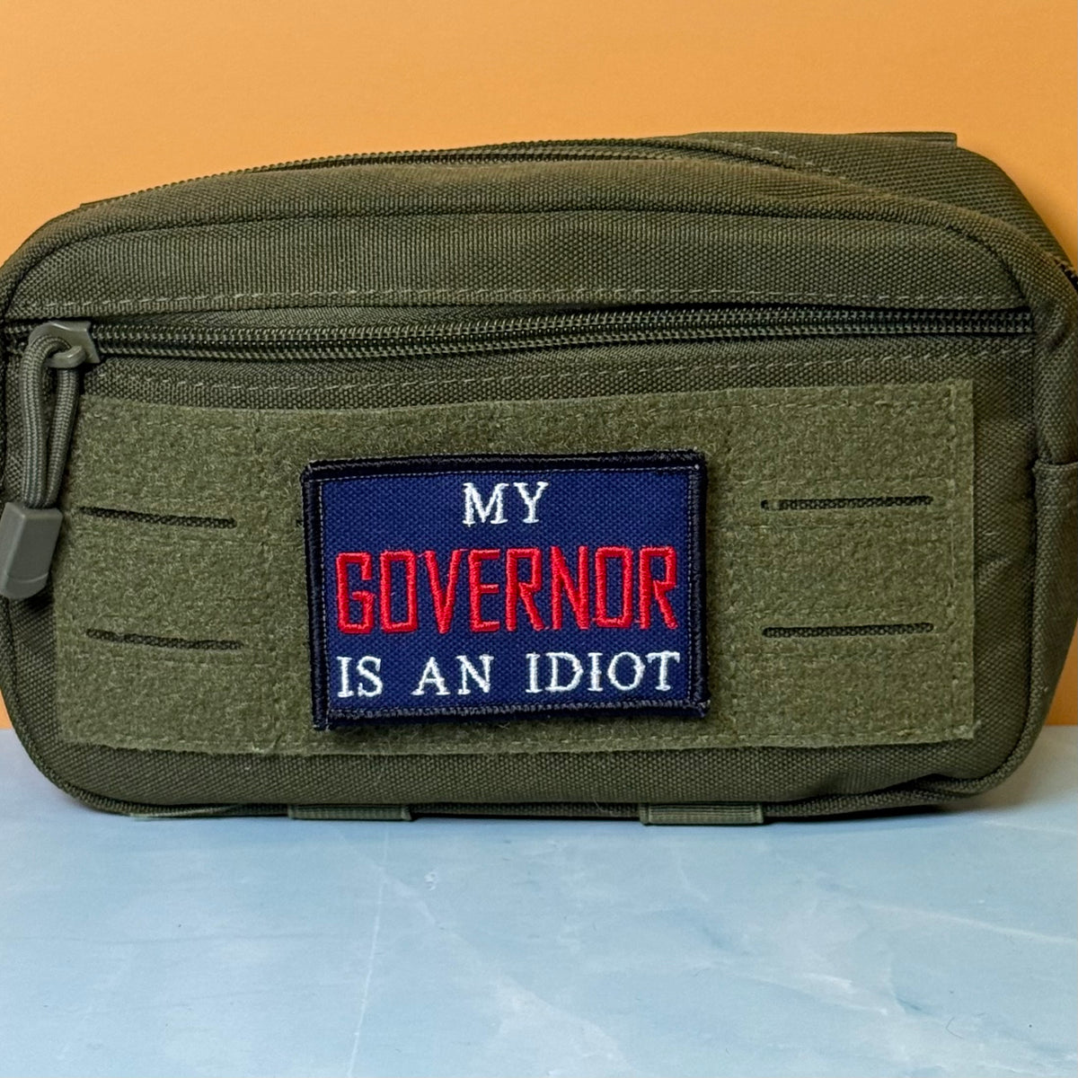 My Governor Is An Idiot embroidered morale patch: olive green tactical pouch with a front zipper and a rectangular embroidered patch that reads 'MY GOVERNOR IS AN IDIOT' in red and white text on a dark blue background, set.