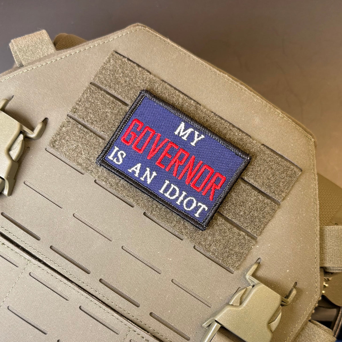 My Governor Is An Idiot embroidered morale patch: with the text 'MY GOVERNOR IS AN IDIOT' in red and white on a blue background, attached to a tan tactical vest with Velcro and plastic buckles.