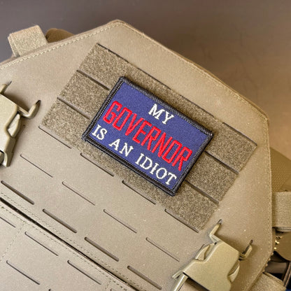 My Governor Is An Idiot embroidered morale patch: with the text 'MY GOVERNOR IS AN IDIOT' in red and white on a blue background, attached to a tan tactical vest with Velcro and plastic buckles.