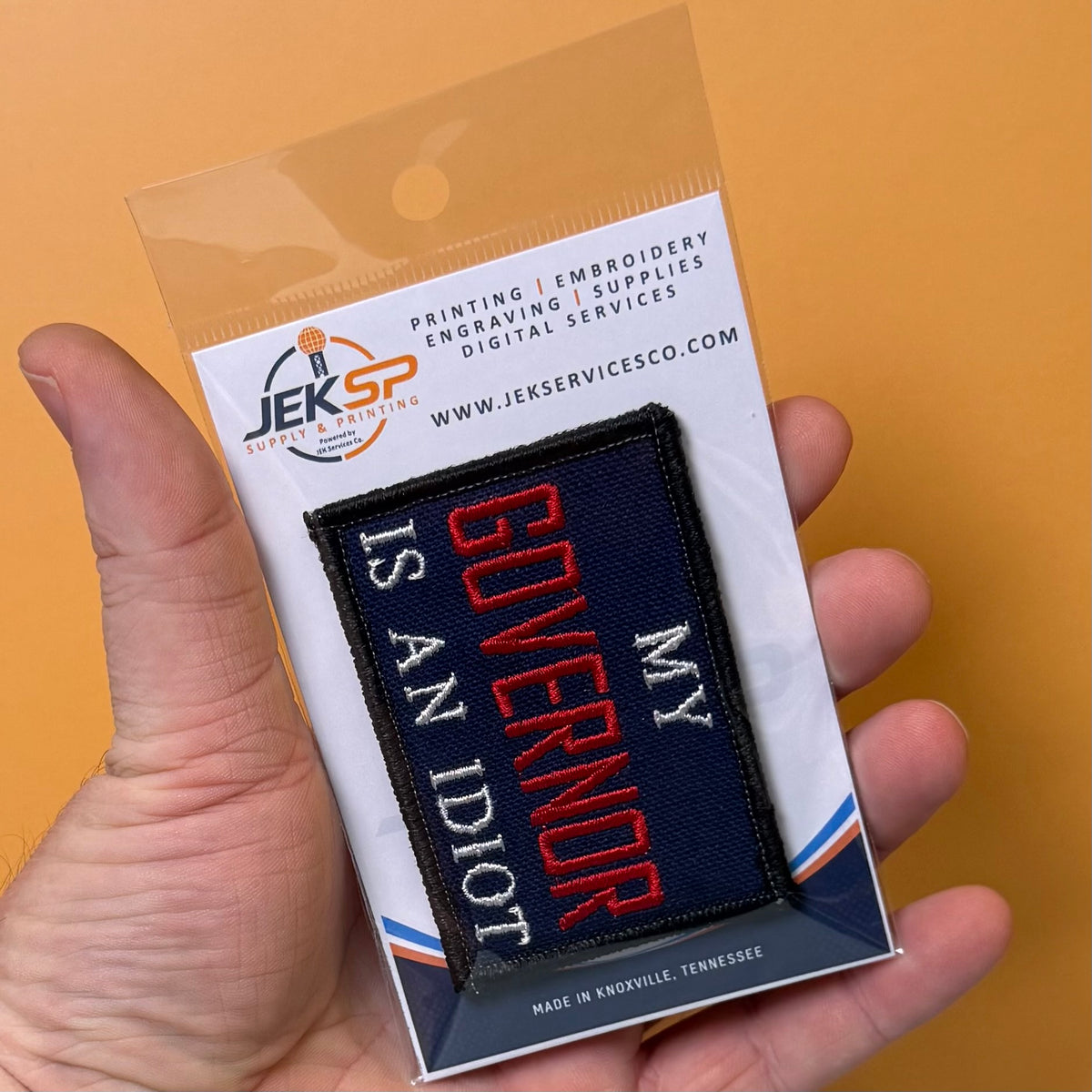 My Governor Is An Idiot embroidered morale patch: hand holding a clear plastic blister pack containing an embroidered patch that reads “MY GOVERNOR IS AN IDIOT.” The package card shows the brand Jeks Services Co.