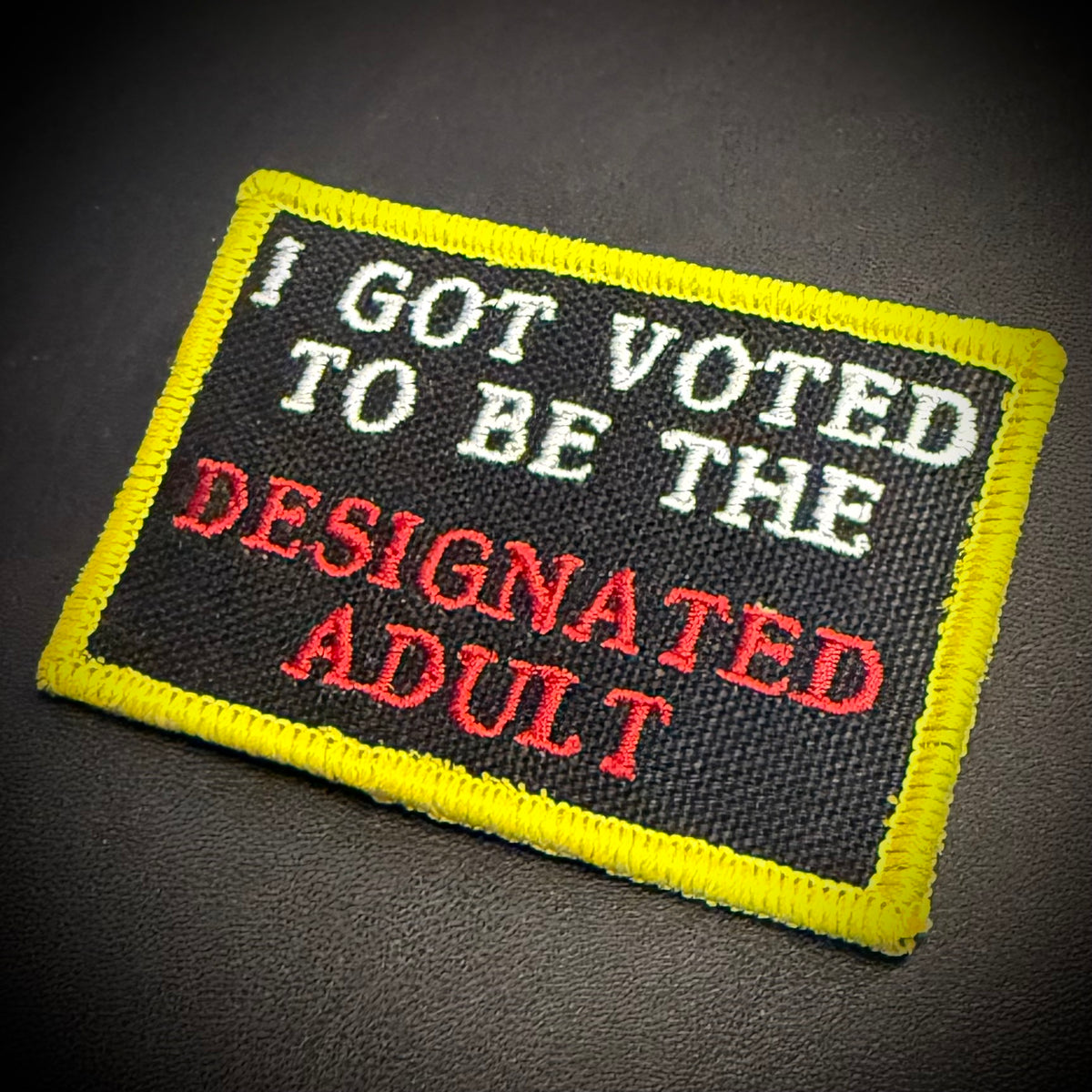 A rectangular embroidered patch with a black background, bright yellow border, and text reading 'I GOT VOTED TO BE THE DESIGNATED ADULT' in white and red lettering, shown on a dark textured surface.