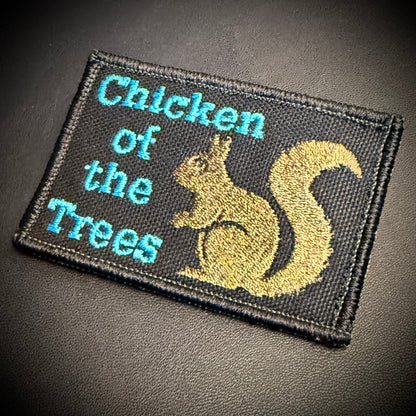 A rectangular embroidered patch with a black background, featuring the text 'Chicken of the Trees' in light blue and a gold embroidered squirrel on the right side, shown on a dark textured surface.