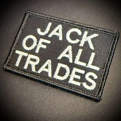 A rectangular black embroidered patch with white text reading 'JACK OF ALL TRADES' on a dark textured background.