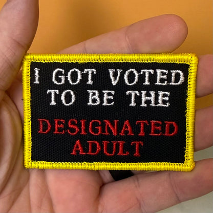 I'm The Designated Adult embroidered morale patch: held in a hand, featuring the text 'I GOT VOTED TO BE THE DESIGNATED ADULT' in white and red embroidery on a black background with a yellow border.