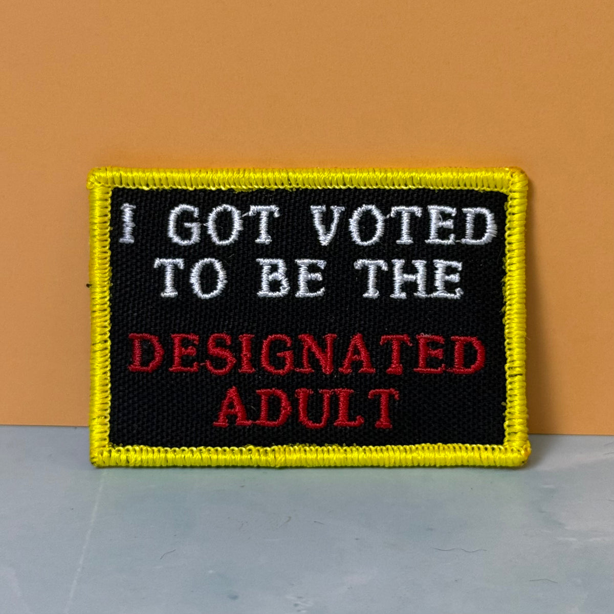 I'm The Designated Adult embroidered morale patch: with a black center, yellow border, white text reading 'I GOT VOTED TO BE THE', and red text reading 'DESIGNATED ADULT', photographed against an orange wall.