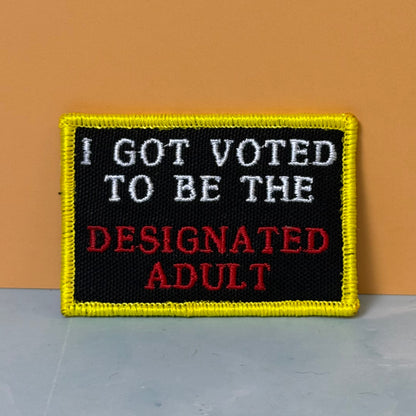 I'm The Designated Adult embroidered morale patch: with a black center, yellow border, white text reading 'I GOT VOTED TO BE THE', and red text reading 'DESIGNATED ADULT', photographed against an orange wall.
