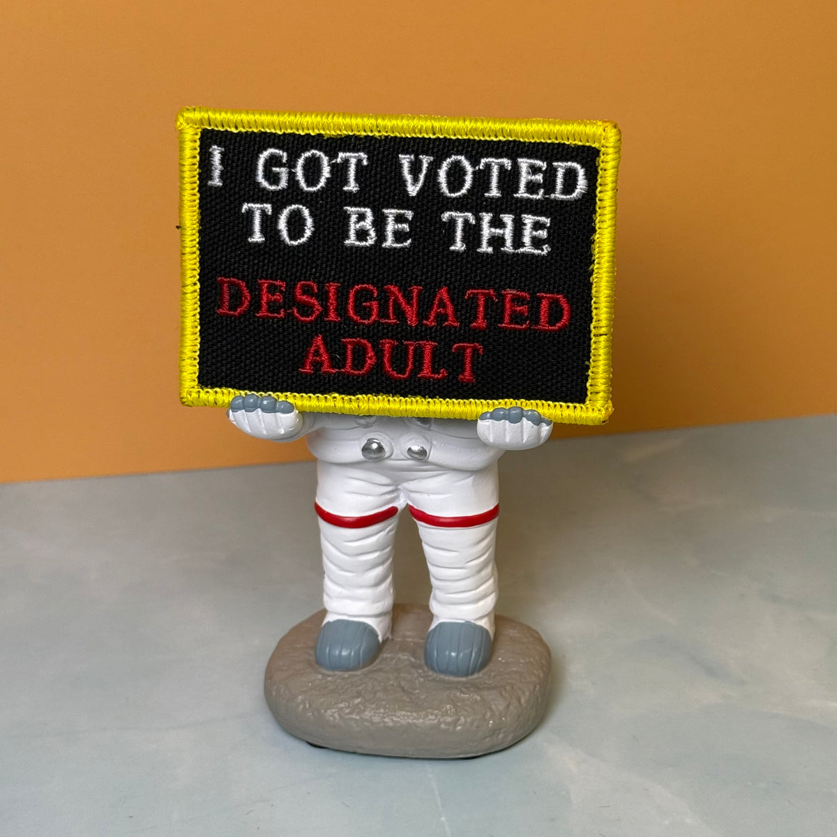 I'm The Designated Adult embroidered morale patch: novelty figurine of an astronaut holding a black sign with the embroidered text 'I GOT VOTED TO BE THE DESIGNATED ADULT' in white and red, set against a solid orange background.