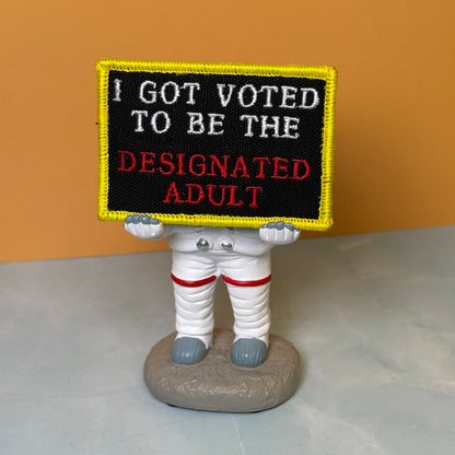I'm The Designated Adult embroidered morale patch: novelty figurine of an astronaut holding a black sign with the embroidered text 'I GOT VOTED TO BE THE DESIGNATED ADULT' in white and red, set against a solid orange background.