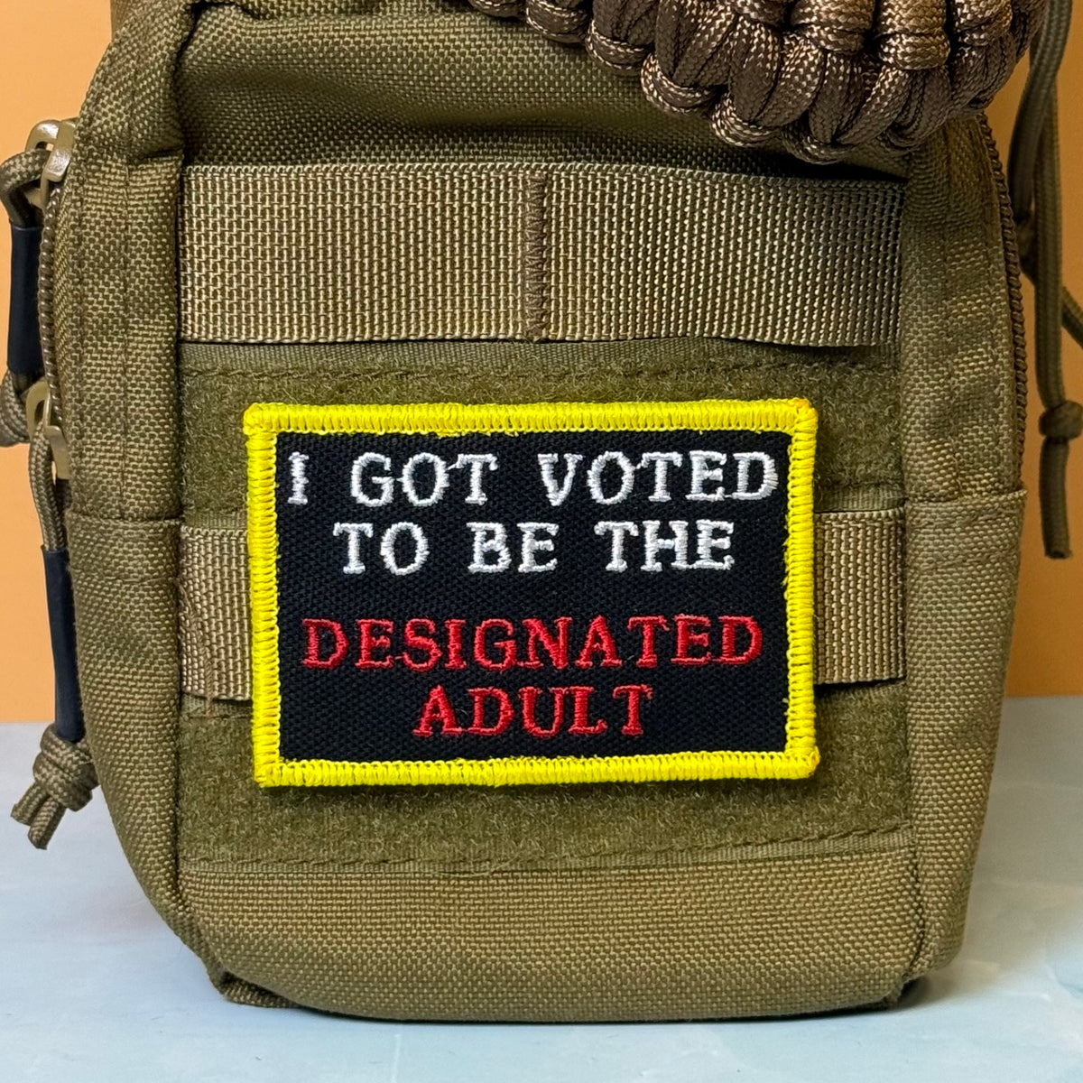 I'm The Designated Adult embroidered morale patch: olive green tactical pouch with a rectangular embroidered patch that reads 'I GOT VOTED TO BE THE DESIGNATED ADULT' in white and red text on a black background with a yellow border.
