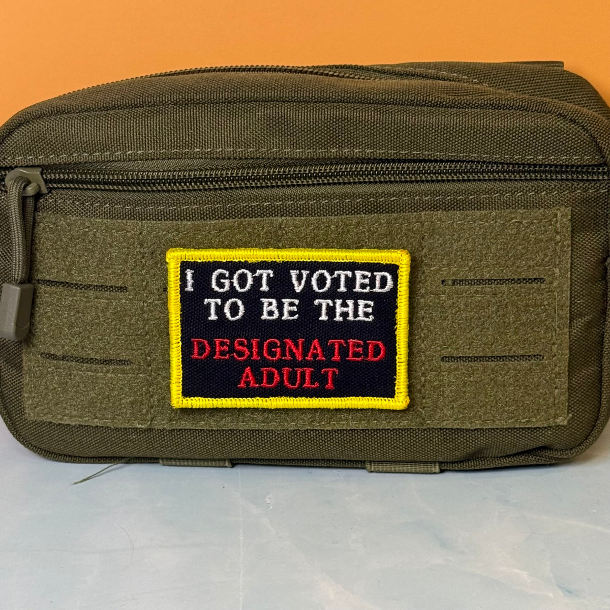 I'm The Designated Adult embroidered morale patch: olive drab tactical pouch with a front hook-and-loop patch that reads 'I GOT VOTED TO BE THE DESIGNATED ADULT' in white and red text on a black background with a yellow border.