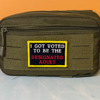 I'm The Designated Adult embroidered morale patch: olive drab tactical pouch with a front hook-and-loop patch that reads 'I GOT VOTED TO BE THE DESIGNATED ADULT' in white and red text on a black background with a yellow border.