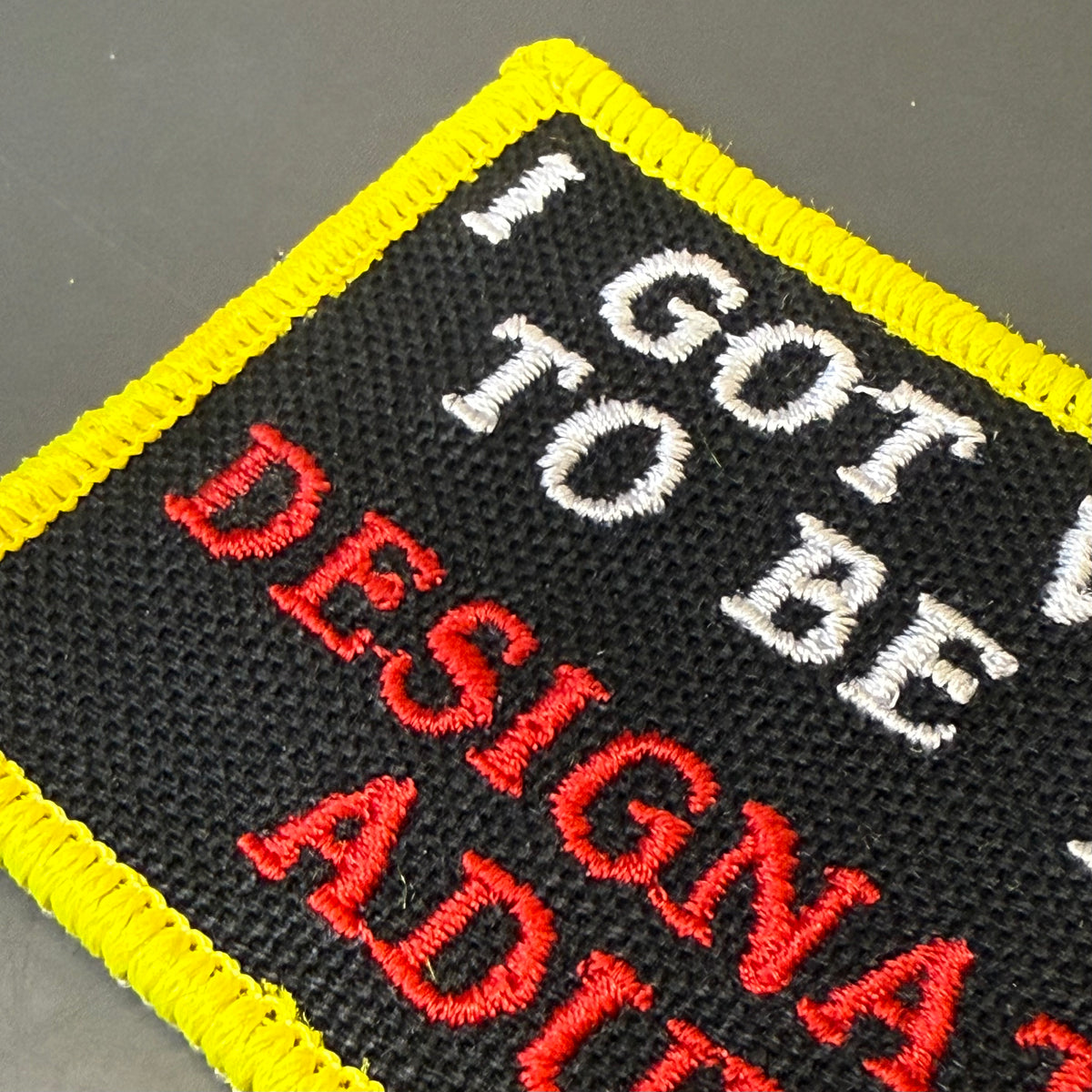 I'm The Designated Adult embroidered morale patch: square embroidered patch with a black background, bright yellow stitched border, and text reading 'I GOT TO BE A DESIGNER' in white and red embroidery, placed on a dark.