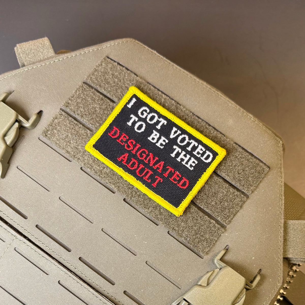 I'm The Designated Adult embroidered morale patch: with a yellow border, black center, and text reading 'I GOT VOTED TO BE THE DESIGNATED ADULT' in white and red, shown attached to tan tactical gear with velcro.