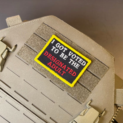 I'm The Designated Adult embroidered morale patch: with a yellow border, black center, and text reading 'I GOT VOTED TO BE THE DESIGNATED ADULT' in white and red, shown attached to tan tactical gear with velcro.
