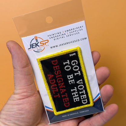 I'm The Designated Adult embroidered morale patch: hand holding a packaged embroidered patch that reads 'I GOT VOTED TO BE THE DESIGNATED ADULT' in white and red text on a black background with a yellow border, against an orange.