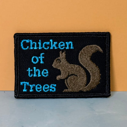 Squirrel, Chicken of the Trees embroidered morale patch: rectangular patch with a black background, blue text reading 'Chicken of the Trees,' and a brown squirrel illustration, shown against a warm orange wall and pale gray surface.