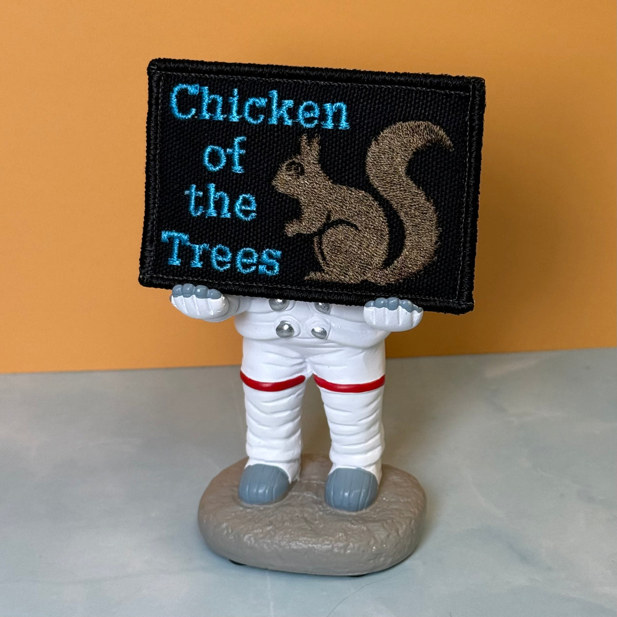 Squirrel, Chicken of the Trees embroidered morale patch: small astronaut figurine holding a black embroidered patch that reads 'Chicken of the Trees' in blue text with a brown squirrel illustration. The astronaut is white.