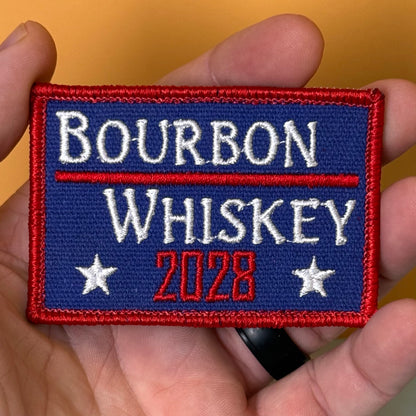 Bourbon & Whiskey for President 2028 embroidered morale patch: held in a hand, featuring the words “BOURBON WHISKEY 2028” in white and red on a blue background with red border and white stars.
