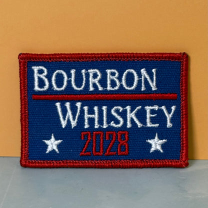 Bourbon & Whiskey for President 2028 embroidered morale patch: with a red border, blue center, white text reading 'BOURBON WHISKEY,' and red and white '2028' with white stars, photographed against a warm tan.