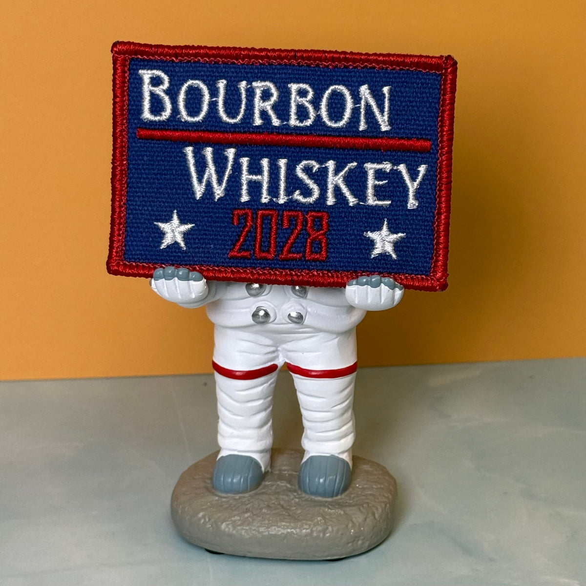 Bourbon & Whiskey for President 2028 embroidered morale patch: small astronaut figurine holding a rectangular embroidered patch that reads “BOURBON WHISKEY 2028” in white and red on a dark blue background. The astronaut is white.