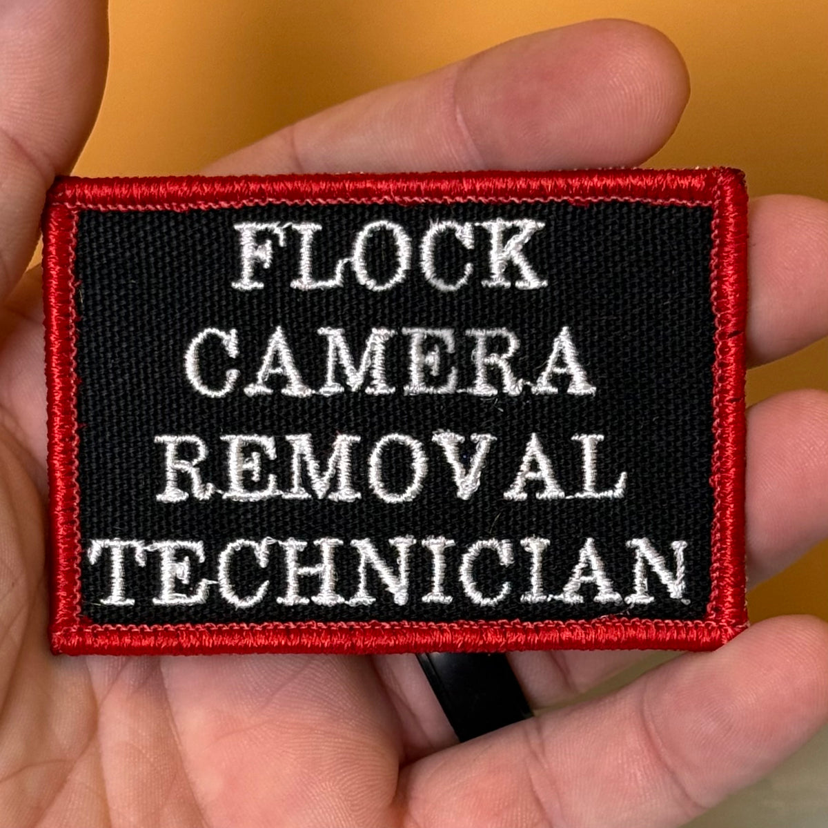 Flock Camera Removal Technician embroidered morale patch: held in a hand, featuring a black background with white text reading 'FLOCK CAMERA REMOVAL TECHNICIAN' and a red stitched border.