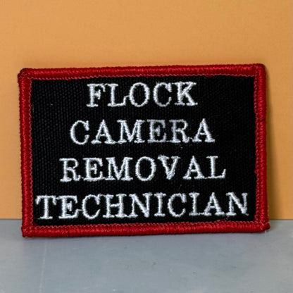 Flock Camera Removal Technician embroidered morale patch: rectangular patch with a black background, red border, and white text reading 'FLOCK CAMERA REMOVAL TECHNICIAN', photographed against a tan wall and gray surface.