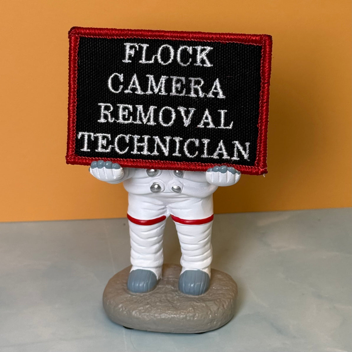 Flock Camera Removal Technician embroidered morale patch: novelty astronaut figurine holding a black rectangular patch with white embroidered text reading 'FLOCK CAMERA REMOVAL TECHNICIAN' and a red border, set against a solid.