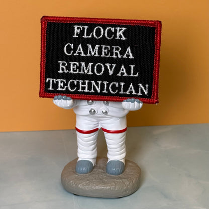 Flock Camera Removal Technician embroidered morale patch: novelty astronaut figurine holding a black rectangular patch with white embroidered text reading 'FLOCK CAMERA REMOVAL TECHNICIAN' and a red border, set against a solid.