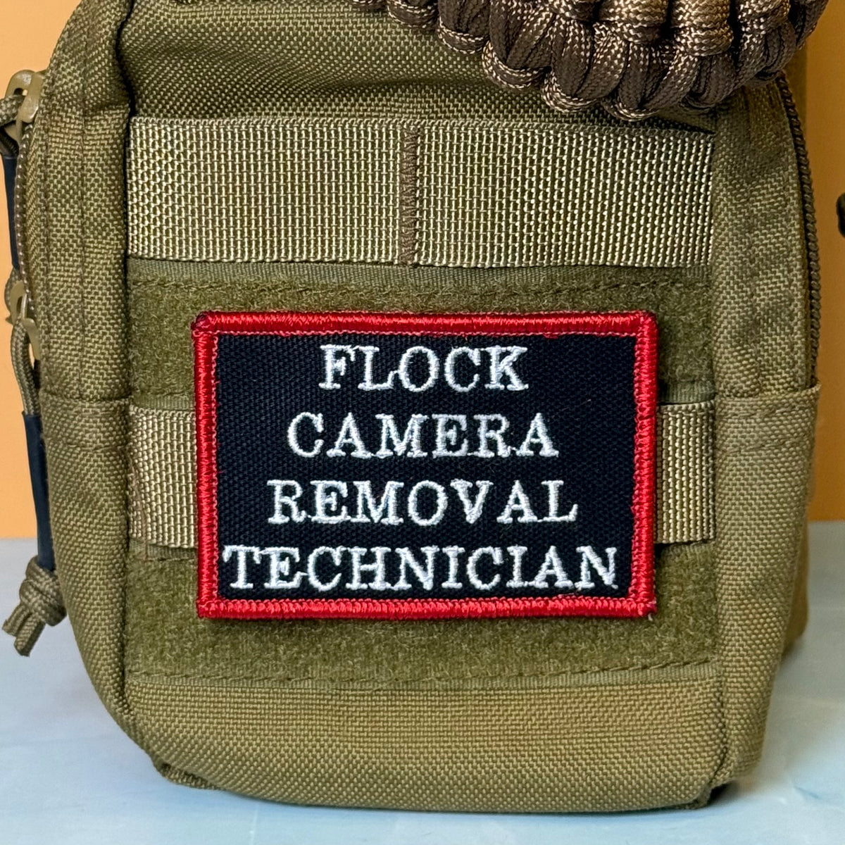 Flock Camera Removal Technician embroidered morale patch: olive green tactical pouch with a black embroidered patch that reads 'FLOCK CAMERA REMOVAL TECHNICIAN' in white text with a red border.