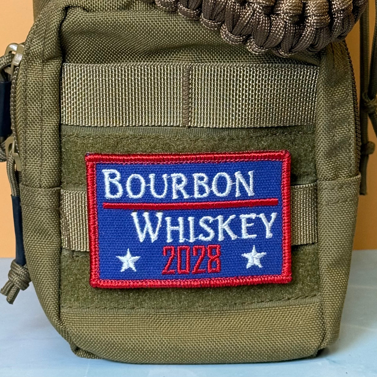 Bourbon & Whiskey for President 2028 embroidered morale patch: olive green tactical pouch featuring a rectangular embroidered patch with a blue background, red border, and white and red text reading 'Bourbon Whiskey 2028'.