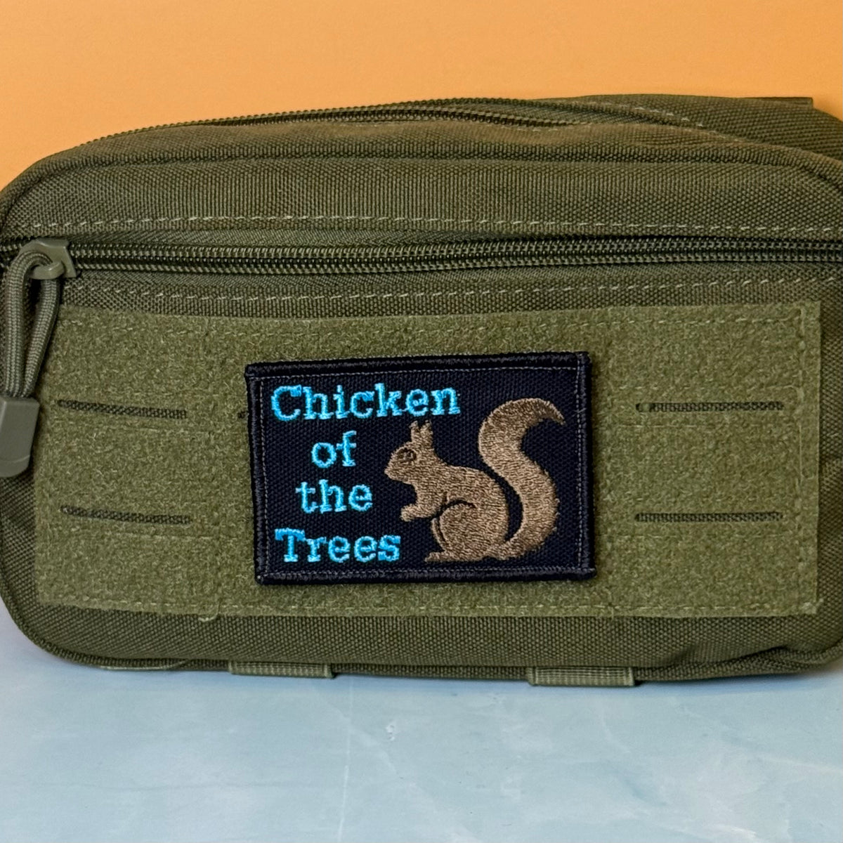 Squirrel, Chicken of the Trees embroidered morale patch: olive green tactical pouch with a front zipper and a rectangular embroidered patch reading 'Chicken of the Trees' in blue text with a brown squirrel design on a black.