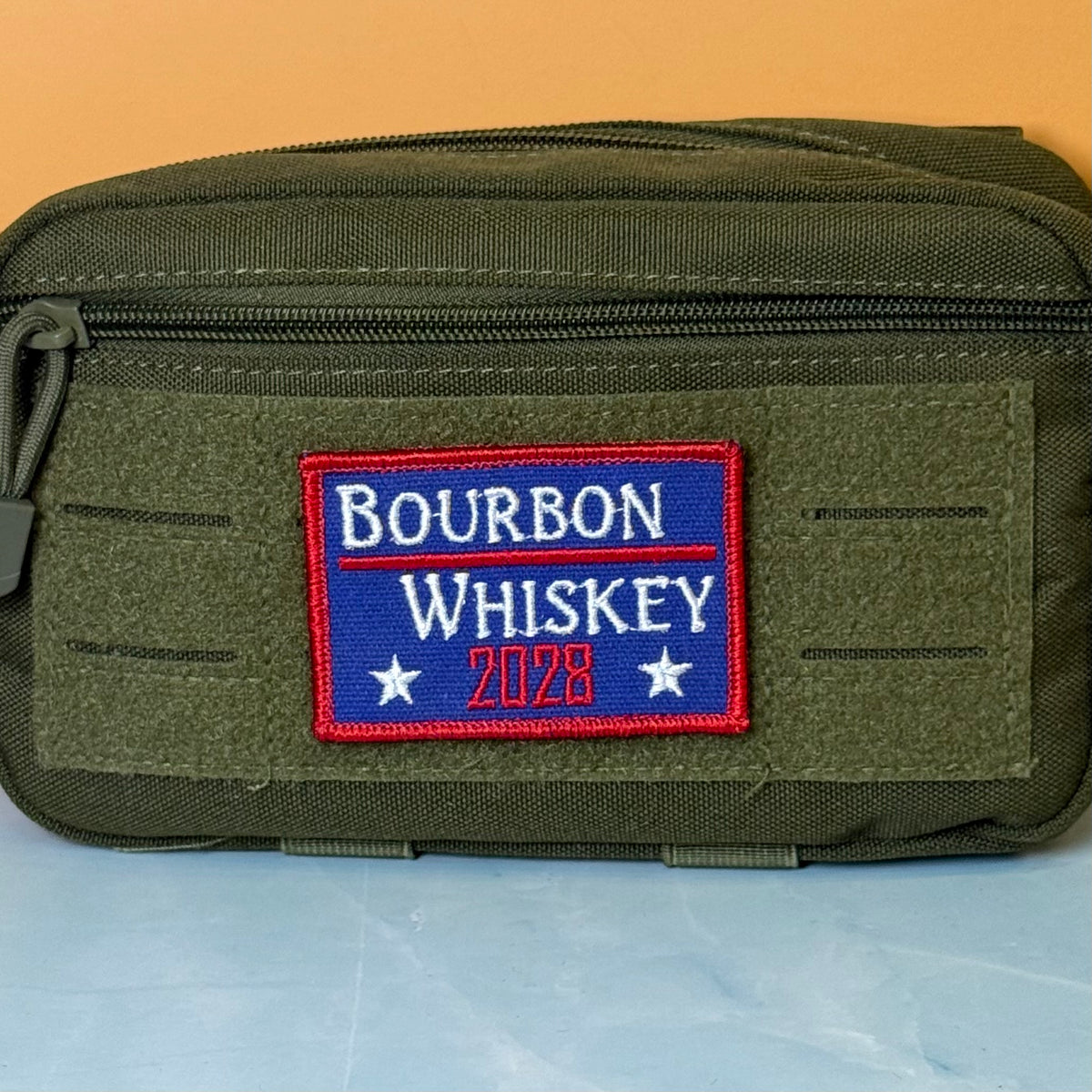 Bourbon & Whiskey for President 2028 embroidered morale patch: olive green tactical pouch featuring a rectangular embroidered patch that reads 'BOURBON WHISKEY 2028' in white and red on a blue background, set against a two-tone.