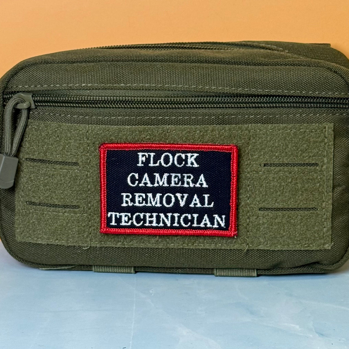 Flock Camera Removal Technician embroidered morale patch: olive green tactical pouch with a front zipper and a rectangular embroidered patch reading 'FLOCK CAMERA REMOVAL TECHNICIAN' in white on a dark navy background with a red.