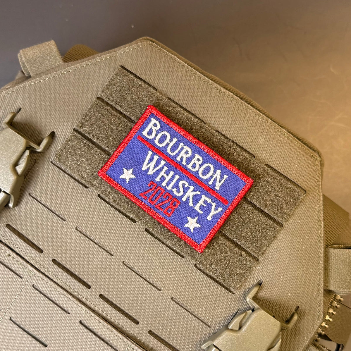 Bourbon & Whiskey for President 2028 embroidered morale patch: reading 'Bourbon Whiskey 2026' in red, white, and blue, attached to tan tactical gear with velcro and buckles.