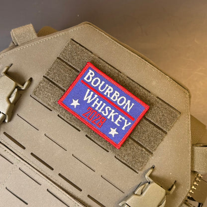 Bourbon & Whiskey for President 2028 embroidered morale patch: reading 'Bourbon Whiskey 2026' in red, white, and blue, attached to tan tactical gear with velcro and buckles.